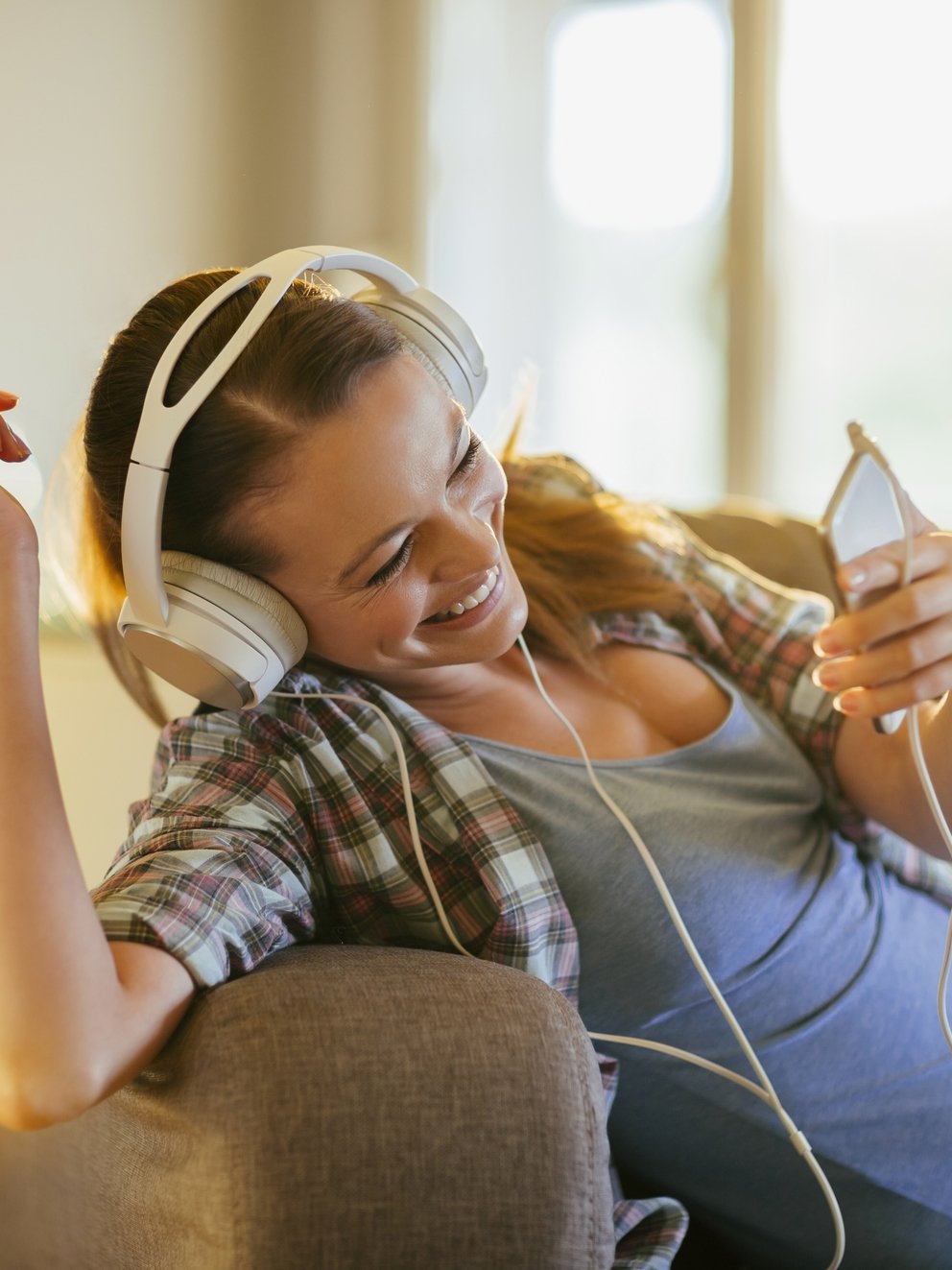 Picture of a young beautiful smiling woman relaxing at home in sofa. She is listening to music threw big headphones. In the background a window is letting in warm sunlight.